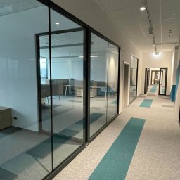 UNITE DK SPÓŁKA Z OGRANICZONĄ ODPOWIEDZIALNOŚCIĄ - Nowoczesny korytarz biurowy z przeszklonymi ścianami działowymi i wykładziną dywanową w odcieniach szarości i turkusu.