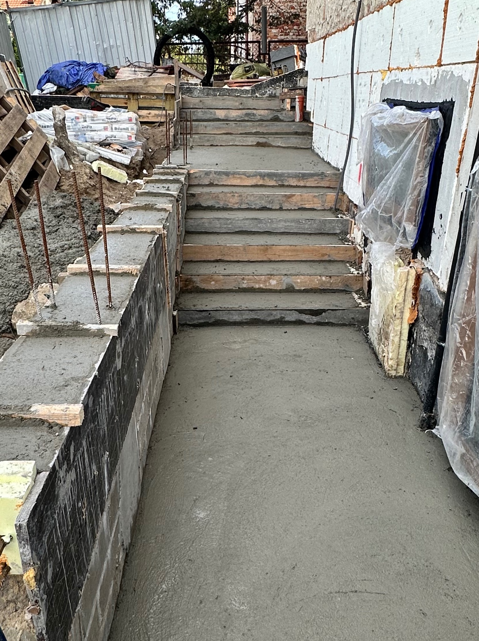Betonowe schody zewnętrzne w trakcie budowy, z widocznymi deskami szalunkowymi i wystającymi prętami zbrojeniowymi, przylegające do ocieplonej ściany budynku.