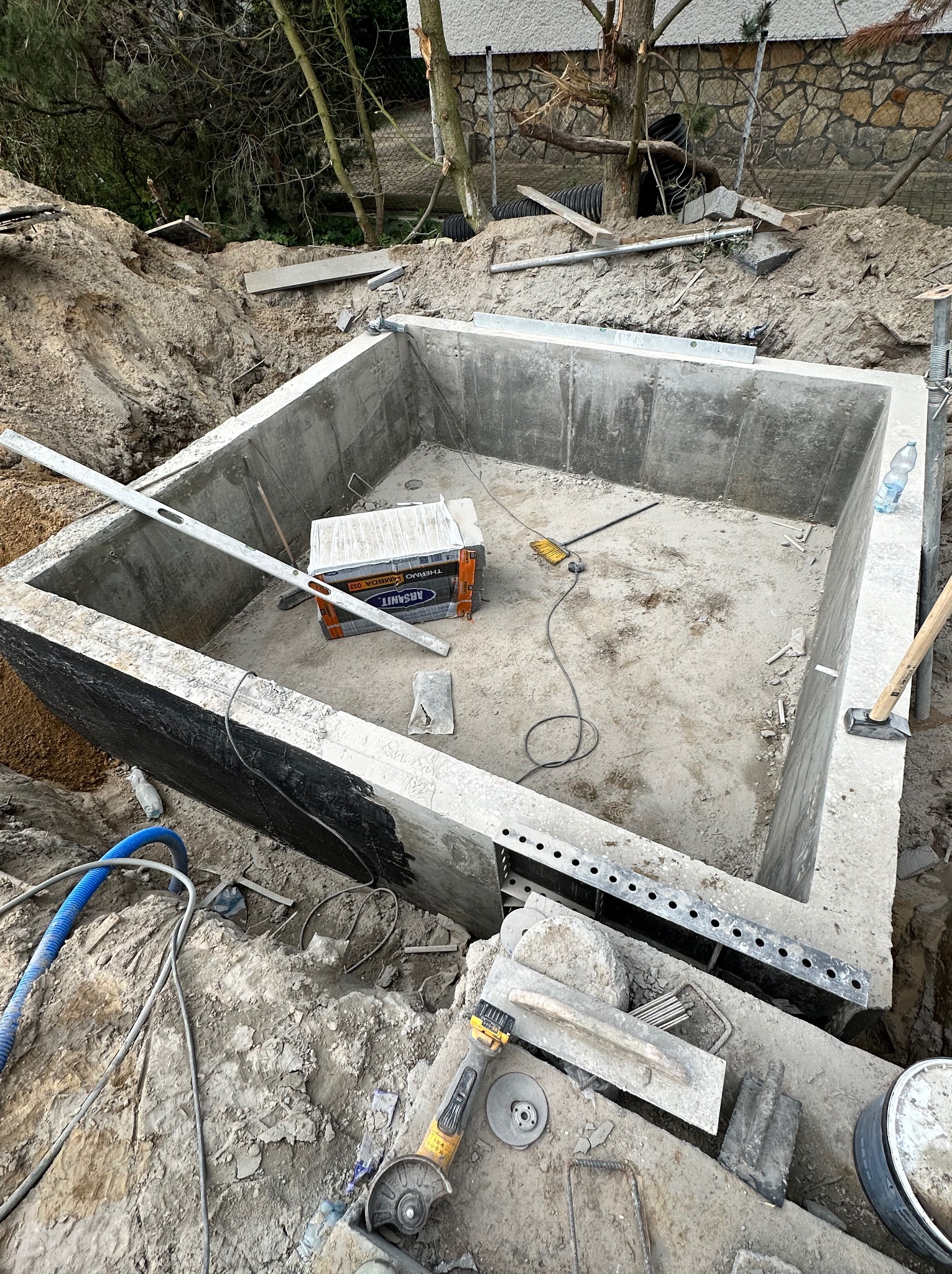 Wykopany dół z betonowymi ścianami w trakcie budowy, widoczne narzędzia budowlane, poziomica, paczka materiałów izolacyjnych, ziemia wokół wykopu.