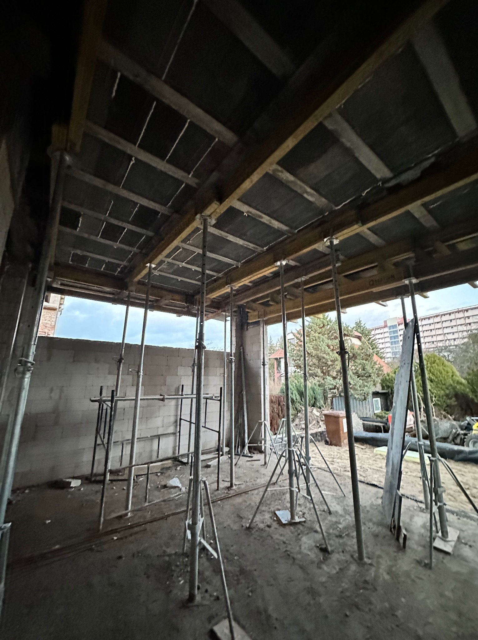 Wnętrze domu w trakcie budowy, widoczne szalunki stropowe wsparte na metalowych podporach, ściany z bloczków betonowych, na zewnątrz widok na budynki mieszkalne i drzewa.