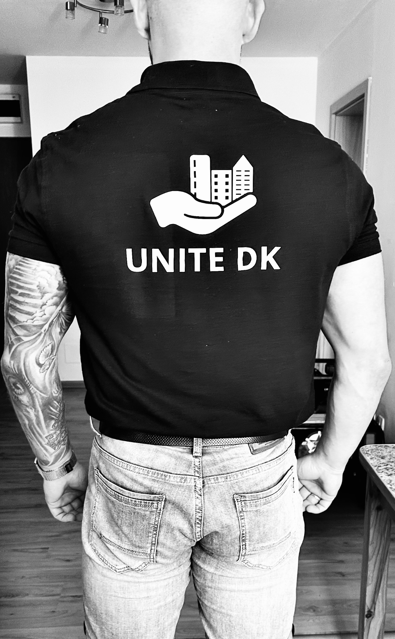 Mężczyzna w czarnej koszulce polo z logo firmy budowlanej Unite DK na plecach, prezentującym dłoń trzymającą budynki, widok od tyłu, z tatuażem na ramieniu.