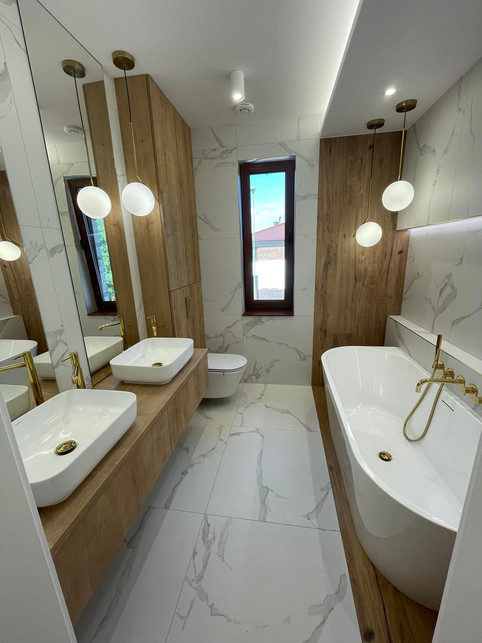 Elegancka łazienka z dwoma umywalkami nablatowymi, złotą armaturą i wanną wolnostojącą, wykończona płytkami imitującymi marmur oraz drewnianymi panelami.