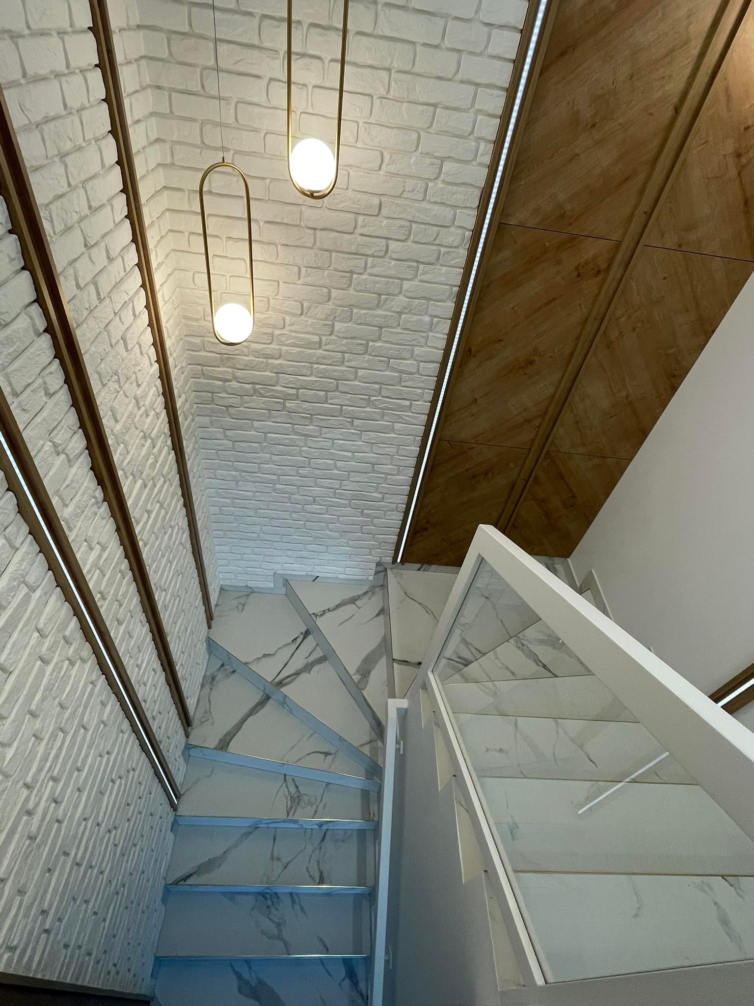 Klatka schodowa z marmurowymi stopniami, szklaną balustradą i oświetleniem LED wzdłuż białej ściany z cegły dekoracyjnej oraz drewnianego sufitu z lampami wiszącymi.
