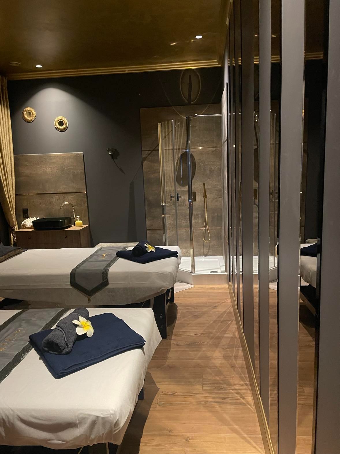 Luksusowe wnętrze gabinetu masażu z dwoma przygotowanymi leżankami, ciemnymi ścianami z dekoracyjnymi elementami, prysznicem w tle i lustrzanymi panelami po prawej stronie.