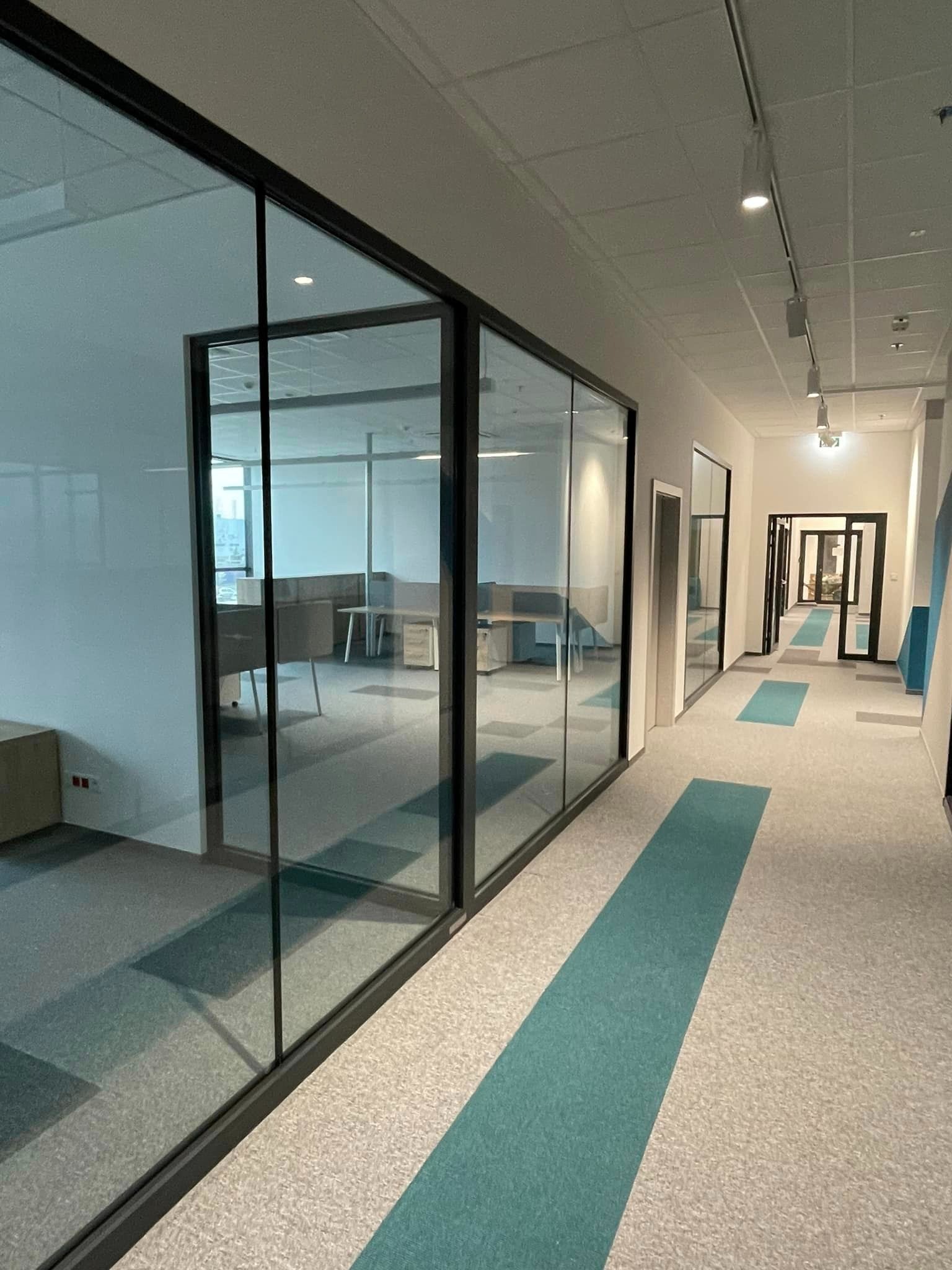 Nowoczesny korytarz biurowy z przeszklonymi ścianami działowymi i wykładziną dywanową w odcieniach szarości i turkusu.