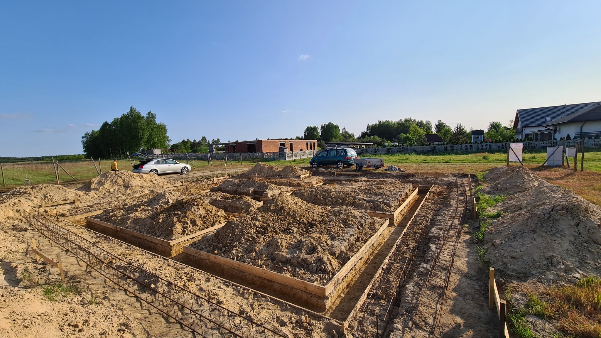 Wykonane deskowanie i zbrojenie pod fundamenty domu jednorodzinnego, widoczne wykopane rowy wypełnione piaskiem, na działce zaparkowane dwa samochody osobowe, w tle zabudowa jednorodzinna.