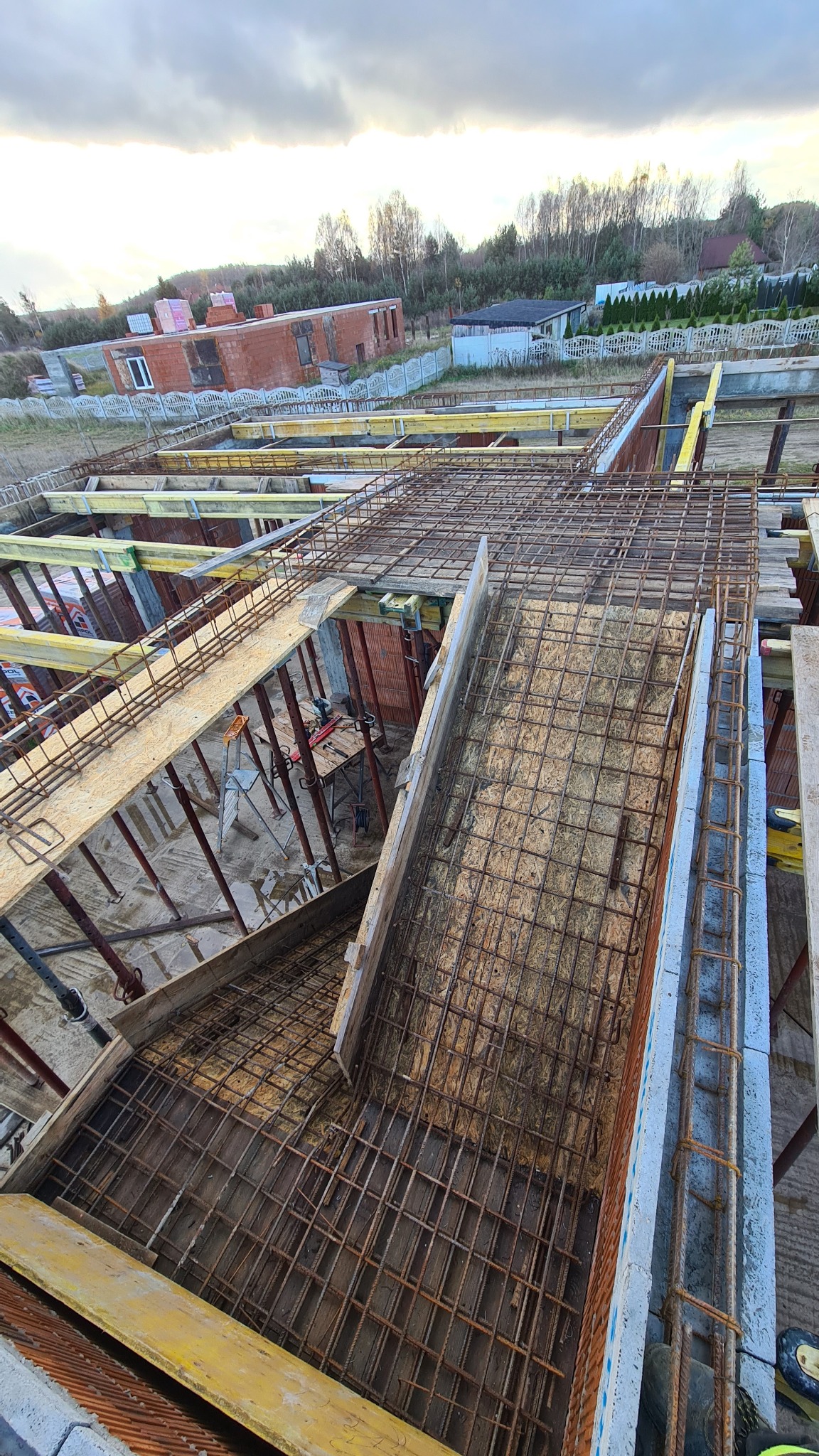 Siatka zbrojeniowa ułożona w szalunkach fundamentów, przygotowana do zalania betonem, widok z góry na plac budowy domu jednorodzinnego w tle.