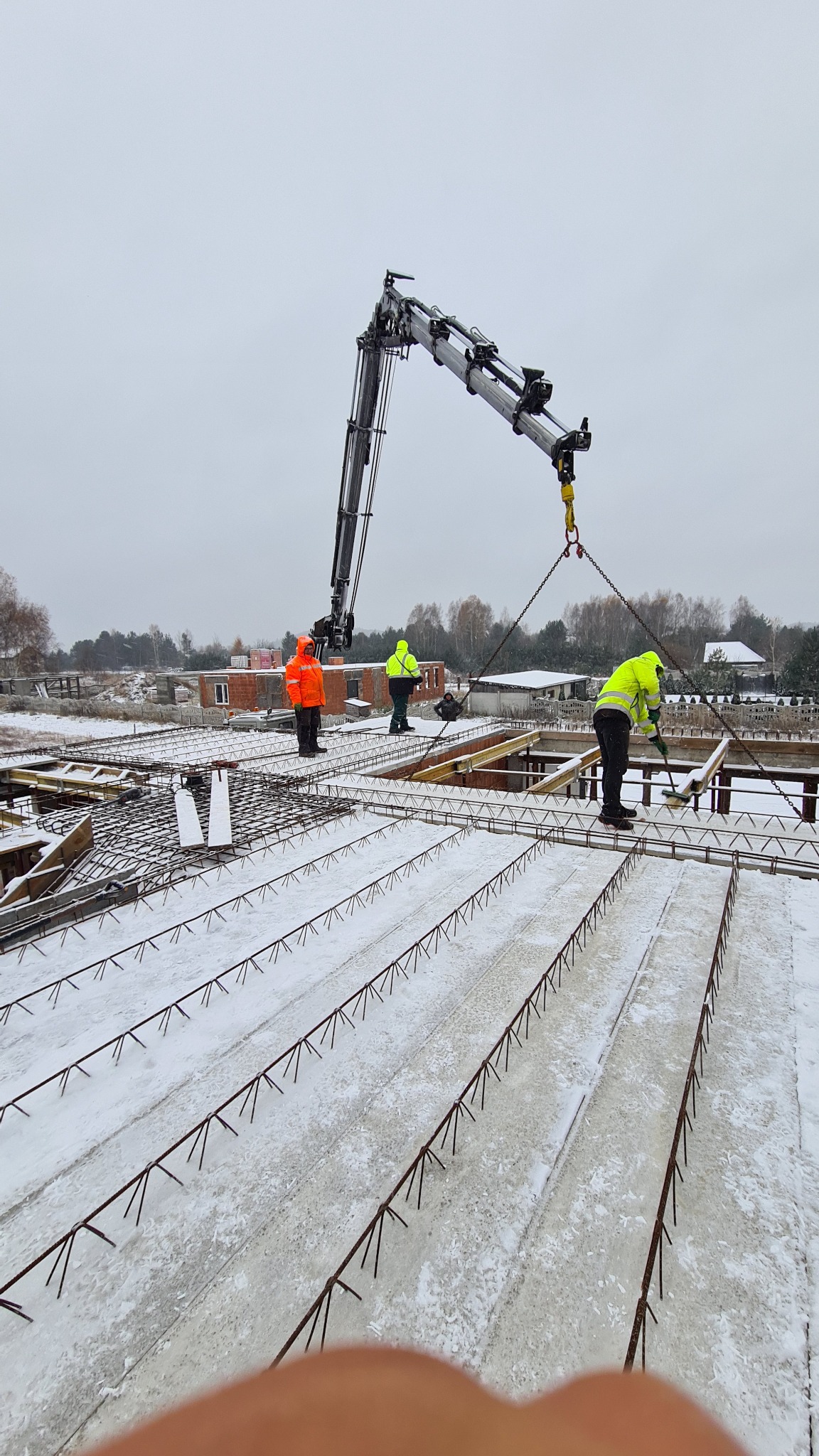 Prace budowlane w zimowych warunkach: robotnicy w odblaskowych ubraniach montują zbrojenie na zaśnieżonym fundamencie, z użyciem dźwigu na tle częściowo wybudowanego domu.