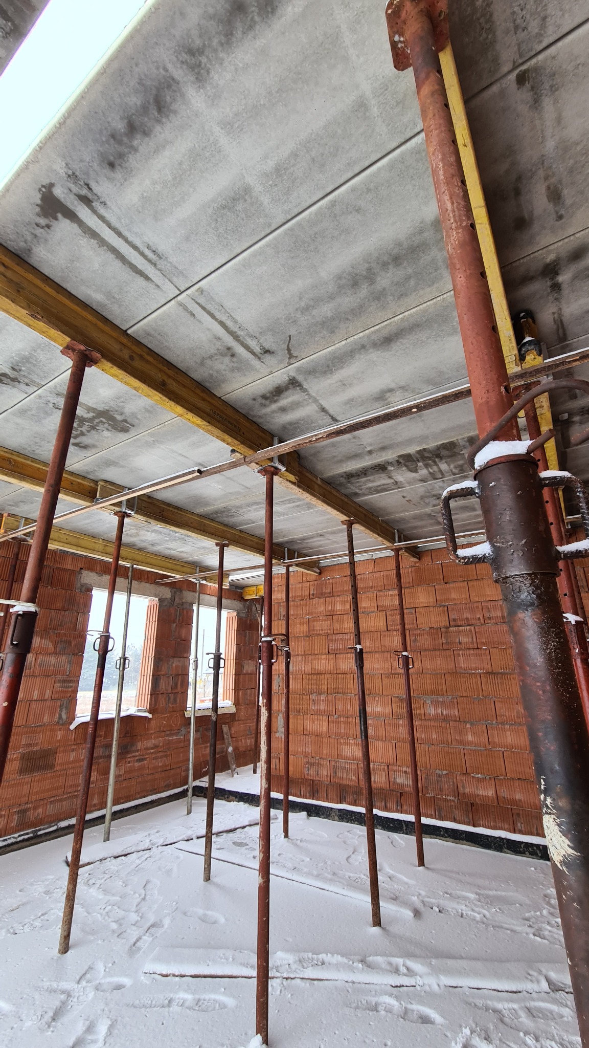 Wnętrze w trakcie budowy domu murowanego, widoczne ściany z czerwonej cegły, tymczasowe podpory stropu pokryte śniegiem, drewniane belki i prefabrykowane płyty stropowe.