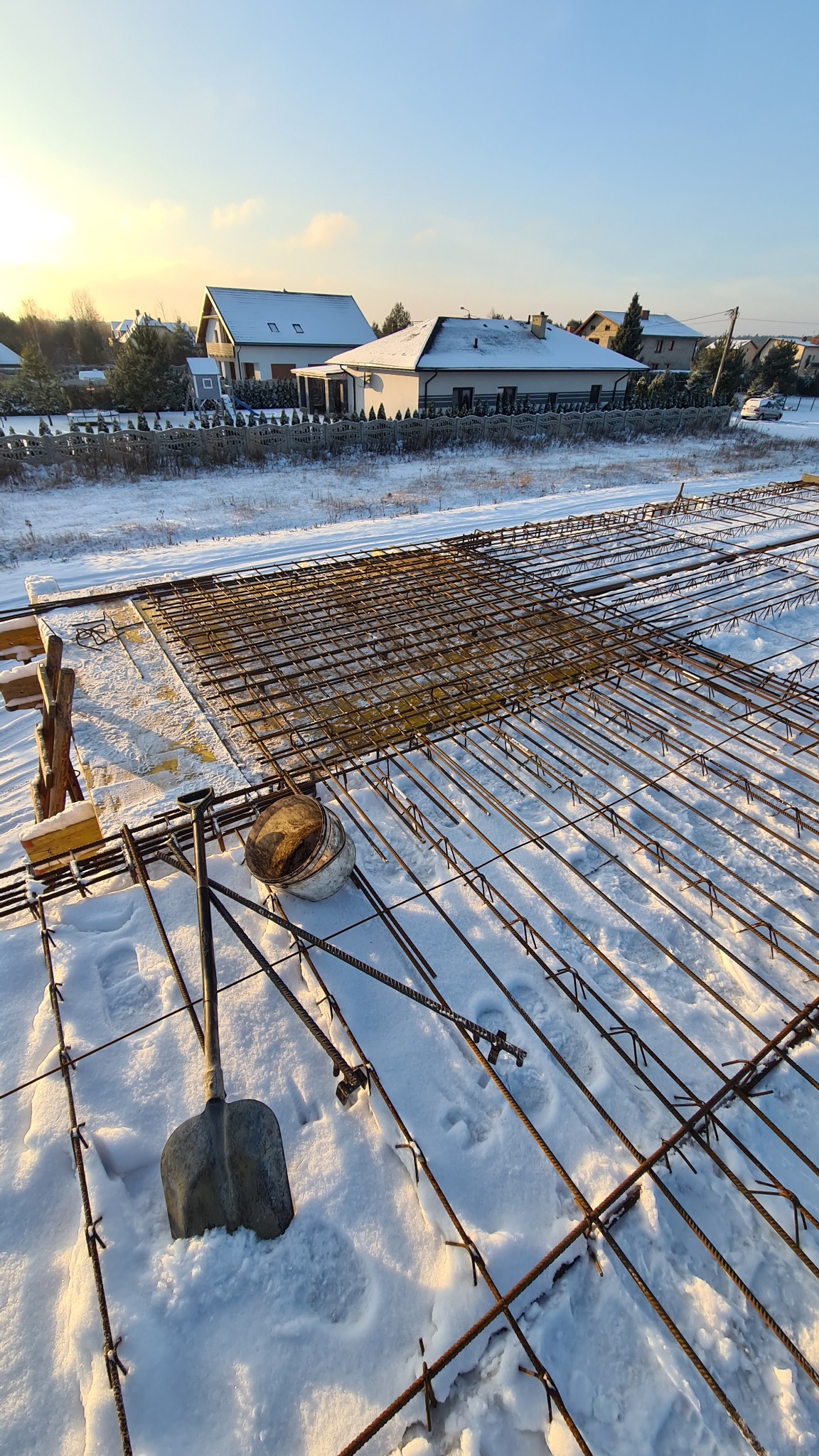 Zimowy widok na plac budowy z ułożonym zbrojeniem fundamentów pokrytym śniegiem, widoczna łopata i wiadro, w tle domy jednorodzinne.