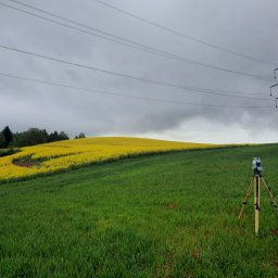 Piotr Wójcik EnergGEO - Tachimetr na trójnogu ustawiony na zielonym polu, w tle wzgórze z kwitnącym rzepakiem i słup wysokiego napięcia pod pochmurnym niebem.