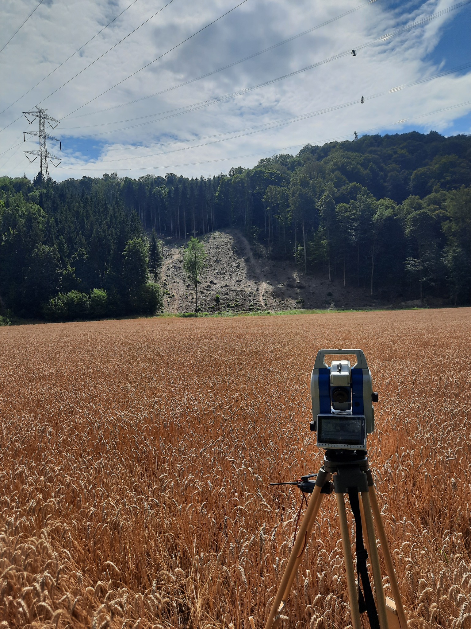 Tachimetr ustawiony na trójnogu na polu zboża, w tle las i linia wysokiego napięcia.