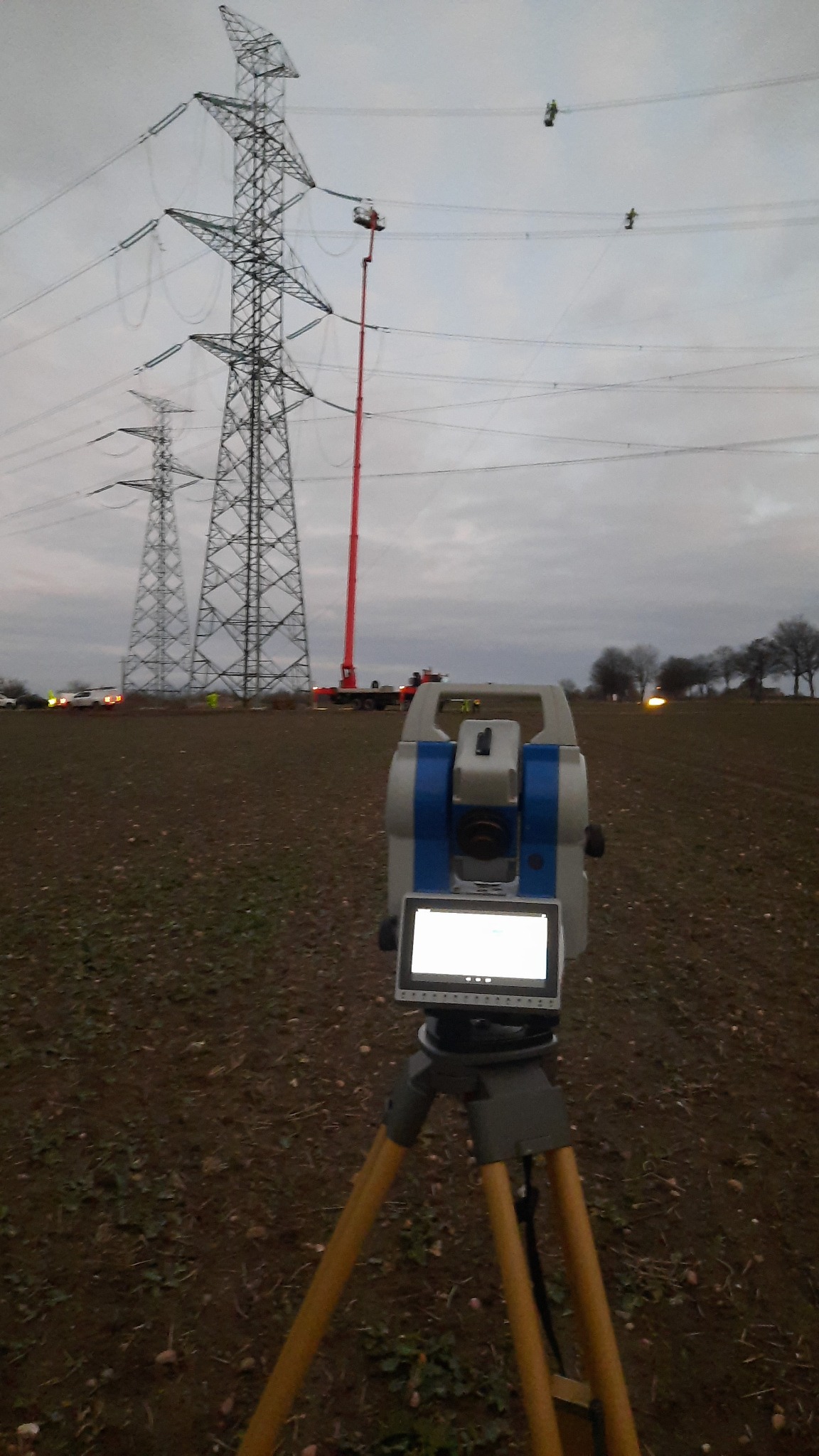 Praca geodezyjna w Gliwicach: Teodolit na trójnogu na polu, w tle słupy wysokiego napięcia z pracownikami na wysokościach, wspierani przez podnośnik koszowy.