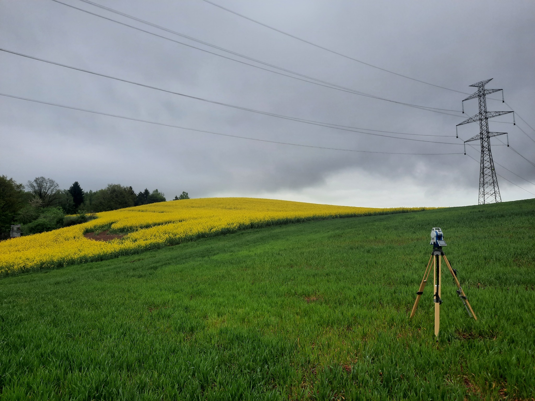 Tachimetr na trójnogu ustawiony na zielonym polu, w tle wzgórze z kwitnącym rzepakiem i słup wysokiego napięcia pod pochmurnym niebem.