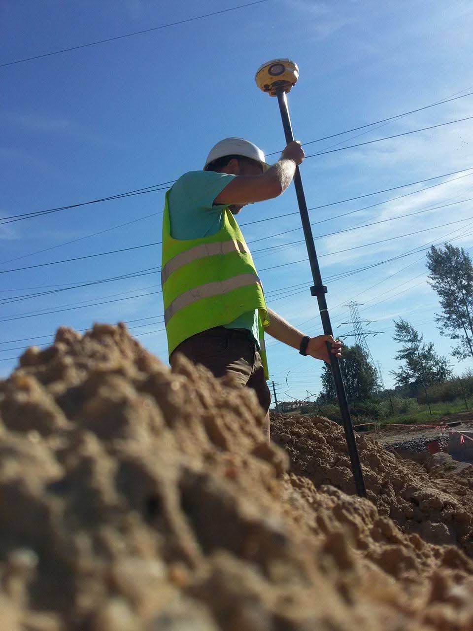 Pracownik w kasku i kamizelce odblaskowej obsługuje geodezyjny odbiornik GPS na tyczce, na tle wykopu i błękitnego nieba z liniami energetycznymi.
