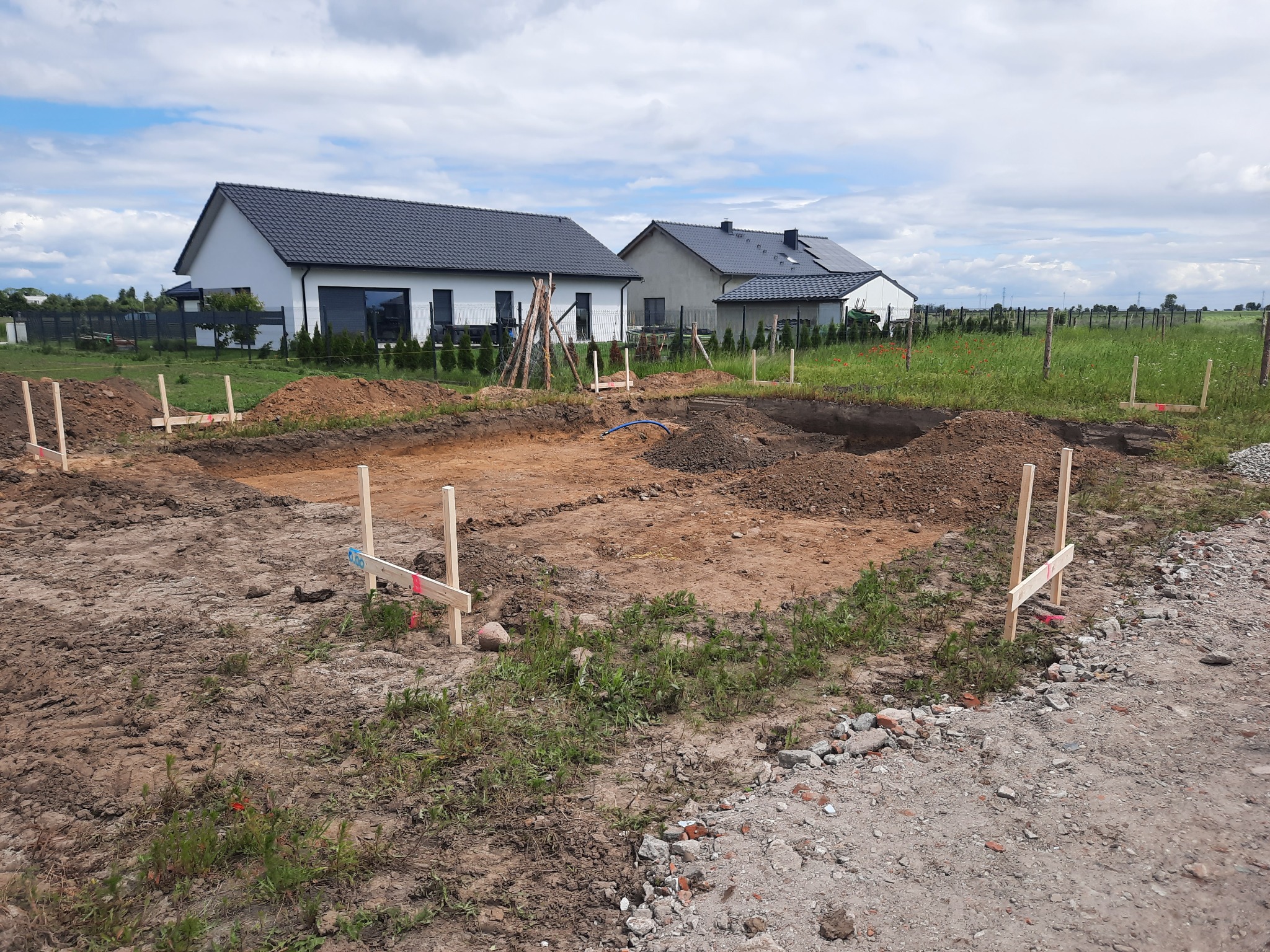 Wytyczanie fundamentów pod budowę domu jednorodzinnego na działce z widokiem na istniejące budynki. Drewniane paliki i wykopane rowy.