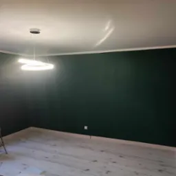 Pomieszczenie po malowaniu ścian na ciemnozielony kolor, widoczna drabina aluminiowa, nowoczesna lampa wisząca i podłoga z jasnych paneli.