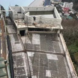 Wysoki kąt widoku na budowę domu w stanie surowym otwartym z widoczną siatką zbrojeniową na dachu, betonowymi ścianami i fragmentem wybrzeża w tle.