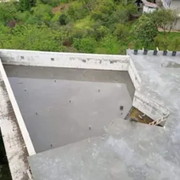 Świeżo wylany beton na dachu budynku z widocznymi elementami konstrukcyjnymi, w tle zieleń drzew i zabudowa mieszkalna.