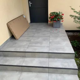 Nowoczesne, szare płytki na schodach wejściowych, karton oparty o ścianę, czarne drzwi wejściowe i donica z czerwonymi kwiatami.