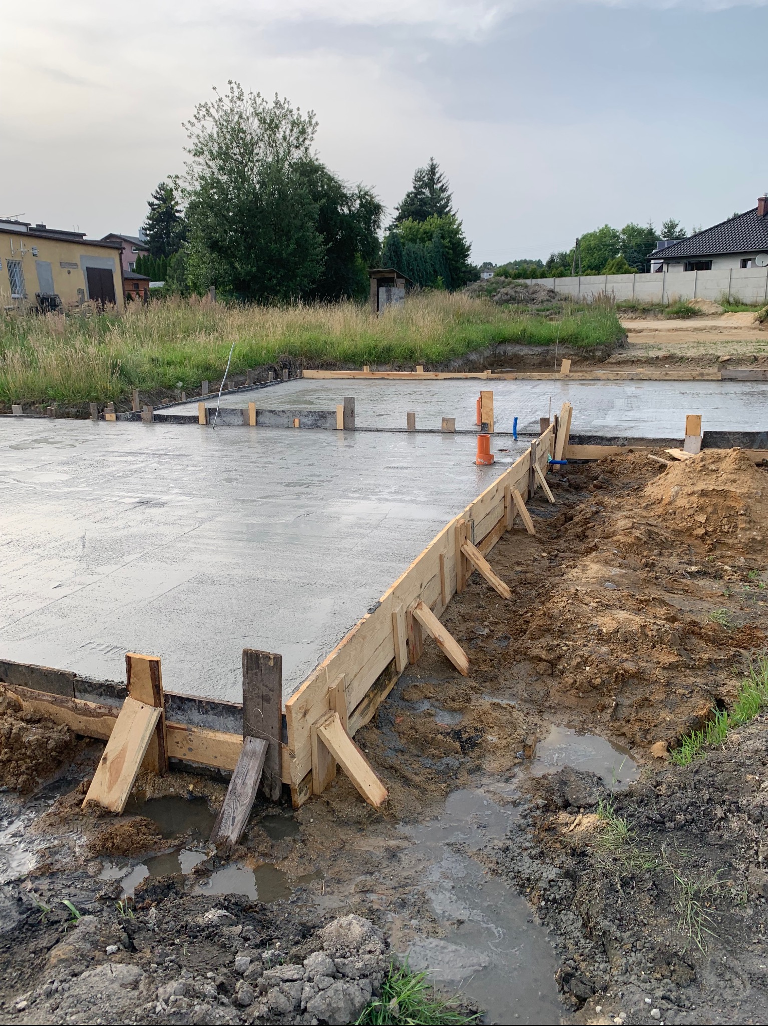 Świeżo wylana, betonowa płyta fundamentowa z drewnianym szalunkiem, widoczne wystające rury instalacyjne, teren budowy z ziemią i kałużami, pochmurne niebo w tle.