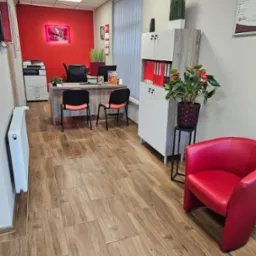 Wnętrze biura z czerwonym akcentem: neon z nazwą firmy nad biurkiem, czerwone krzesło i rośliny doniczkowe, na podłodze płytki imitujące drewno.