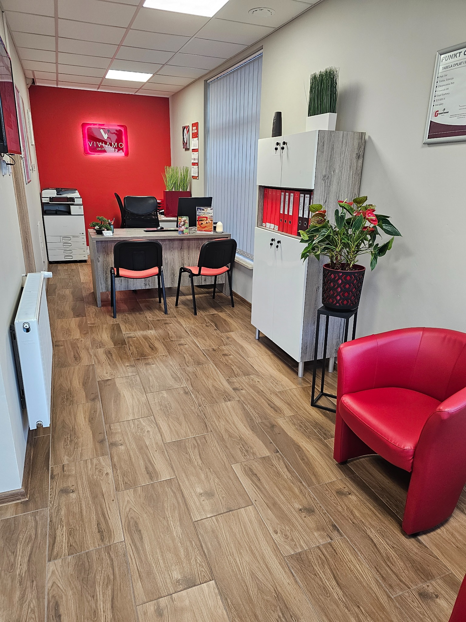 Wnętrze biura z czerwonym akcentem: neon z nazwą firmy nad biurkiem, czerwone krzesło i rośliny doniczkowe, na podłodze płytki imitujące drewno.