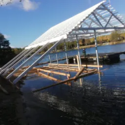 Konstrukcja z białych belek i drewnianych bali wystająca z wody jeziora, widoczny szkielet dachu i podstawa, w tle pomost i drzewa.
