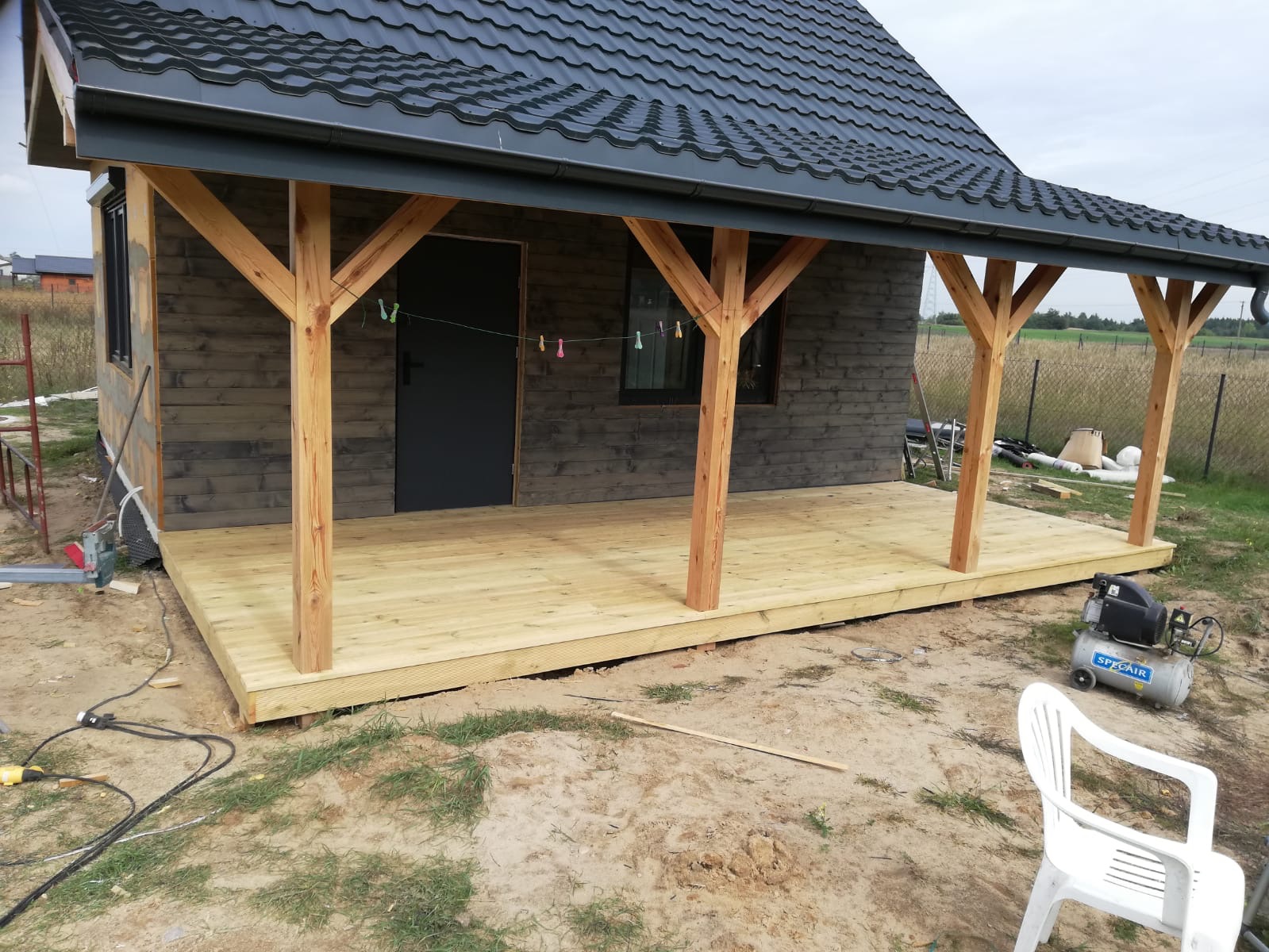 Surowy, drewniany budynek z zadaszonym tarasem na etapie budowy; widoczne narzędzia, kompresor i plastikowe krzesło na piaszczystym gruncie.