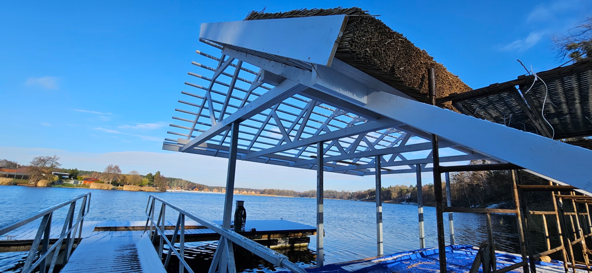 Konstrukcja altany z trzcinowym dachem w trakcie budowy, widok z pomostu nad jeziorem w słoneczny dzień.