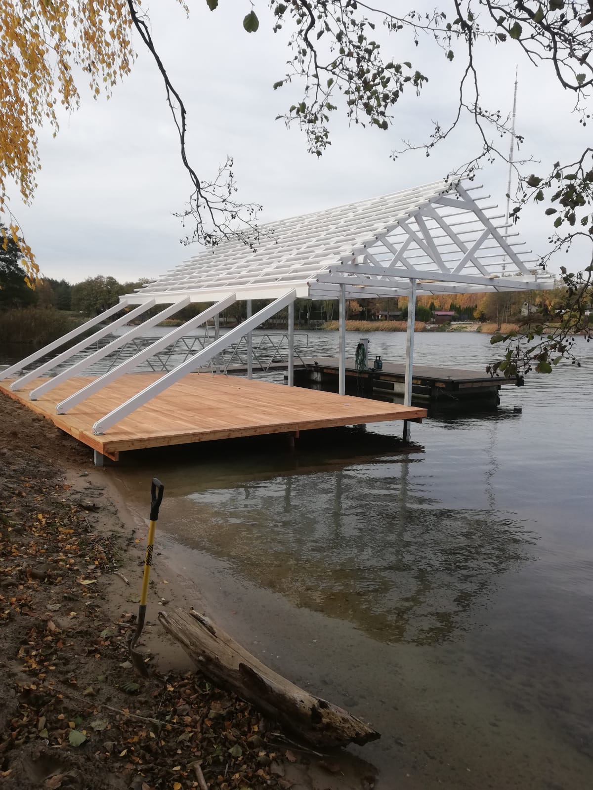 Wczesna faza budowy drewnianego pomostu z białą konstrukcją zadaszenia nad wodą, widoczna łopata na brzegu.