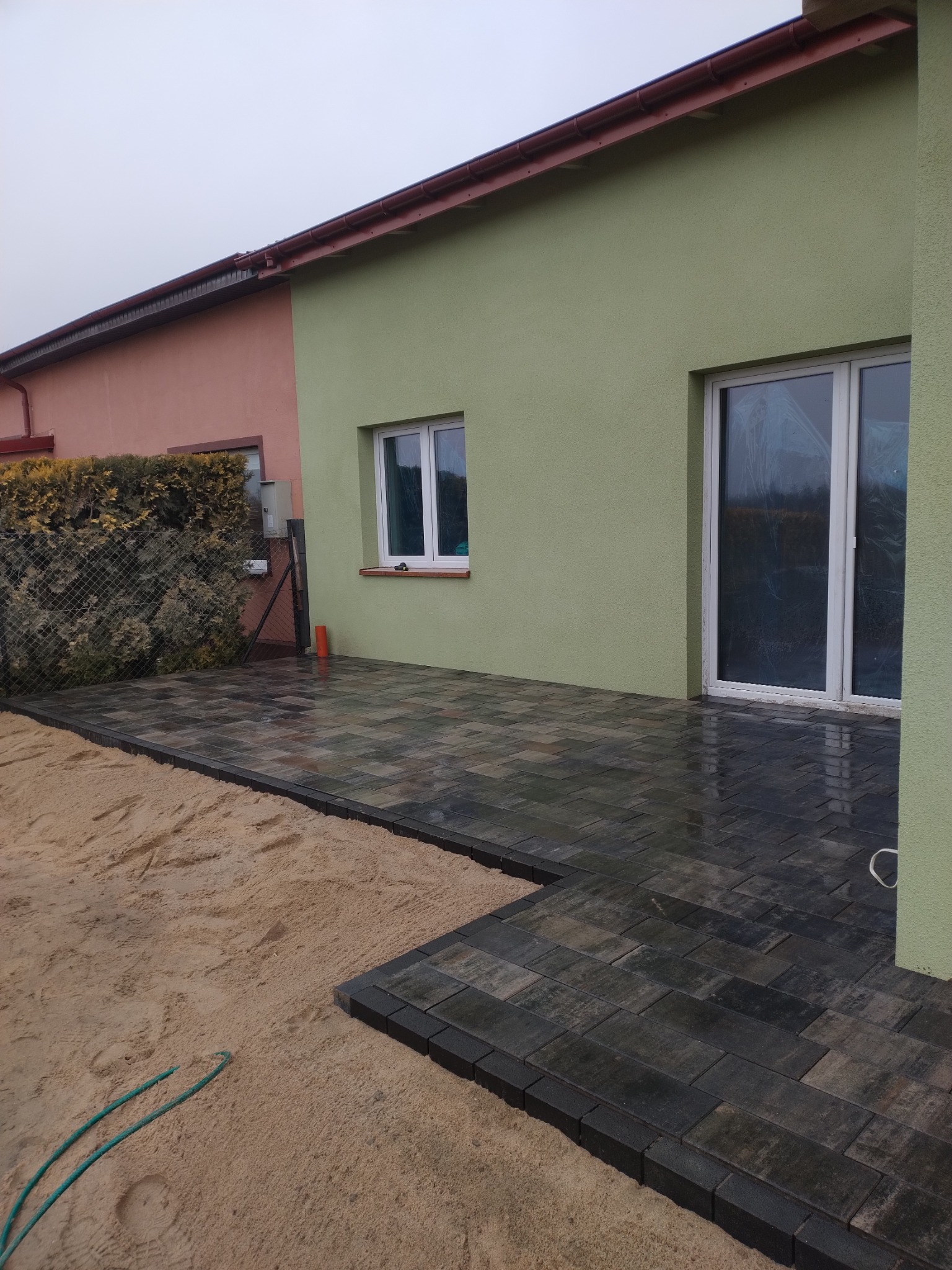 Ułożona, ciemnoszara kostka brukowa na podwórku przylegającym do domu o zielonej elewacji. Widoczny fragment piasku z wężem ogrodowym i okno z folią ochronną.