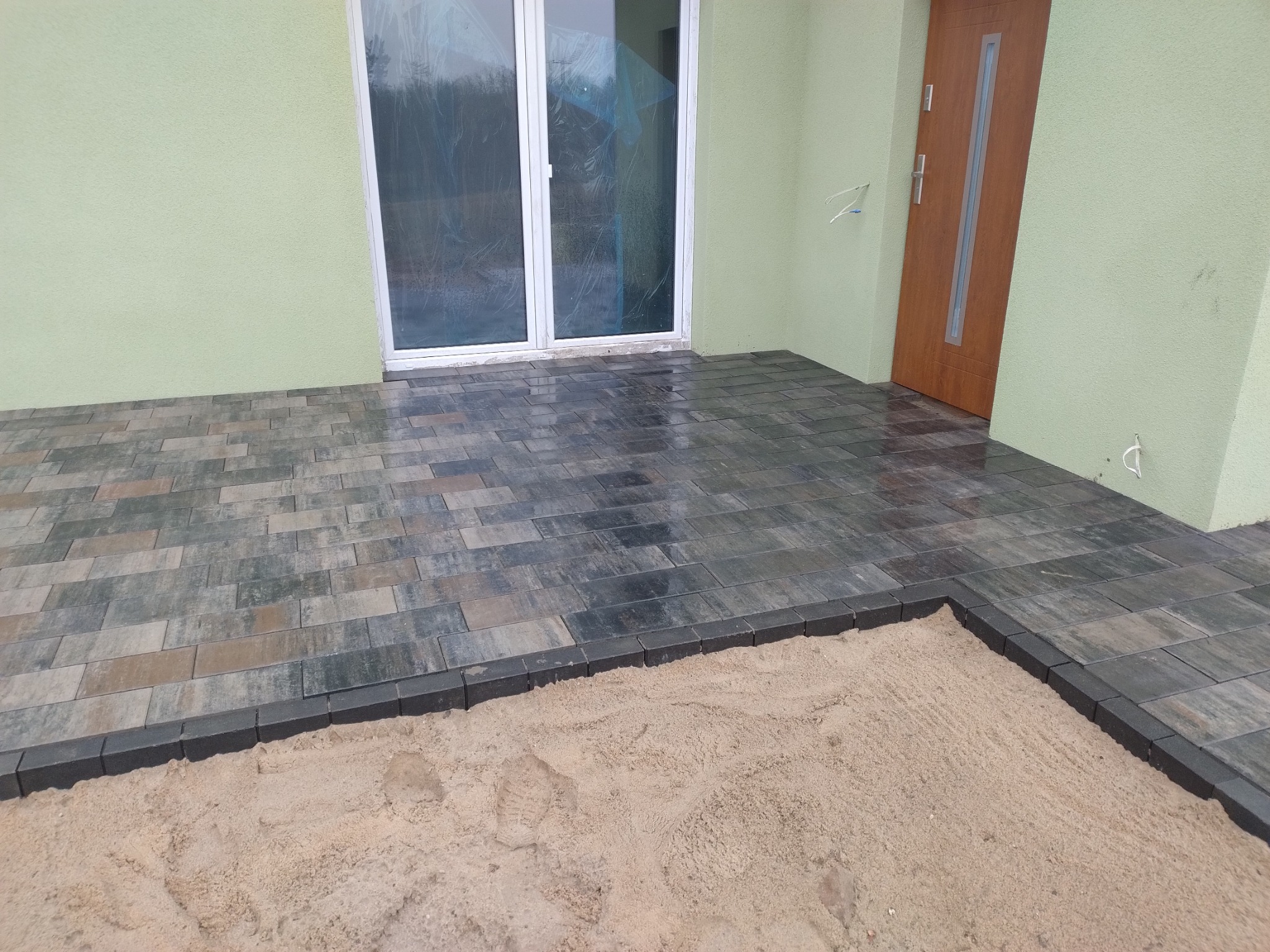 Ułożona kostka brukowa przed domem z zieloną elewacją, widoczne niedokończone obrzeże z piaskiem i ślad buta.