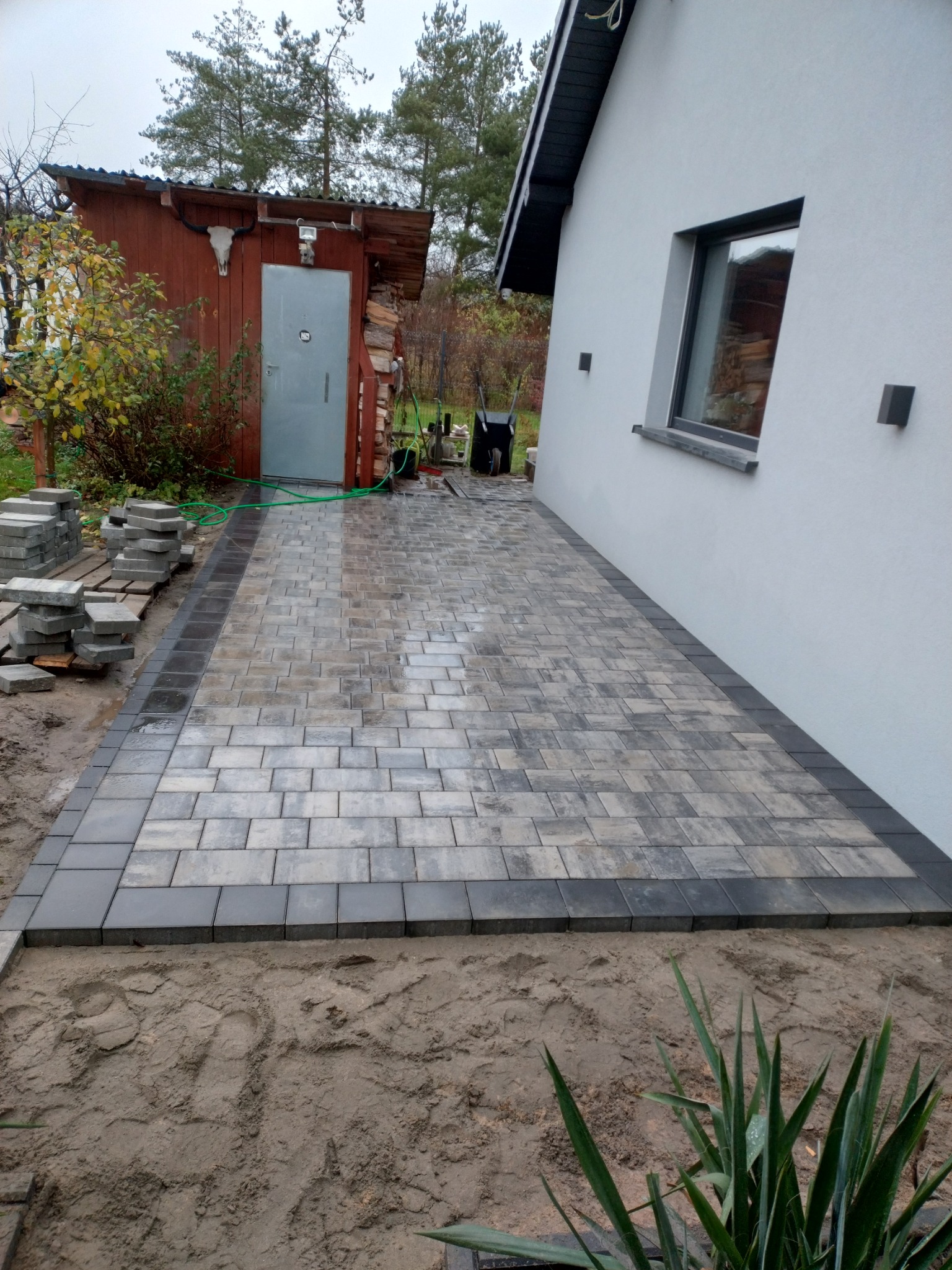 Ułożona ścieżka z kostki brukowej w odcieniach szarości, obramowana ciemniejszą kostką, prowadząca od budynku z oknem do drewnianej szopy z czaszką zwierzęcia na ścianie, widoczne stosy kostki...