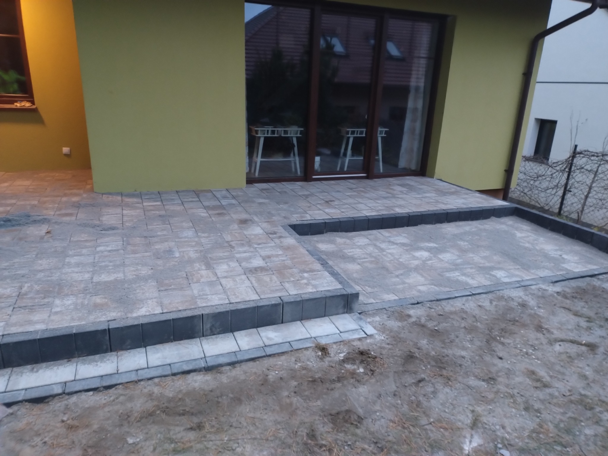 Niedokończony taras z kostki brukowej przy domu o żółtej elewacji, widoczne obrzeża i fragmenty nieułożonej powierzchni.