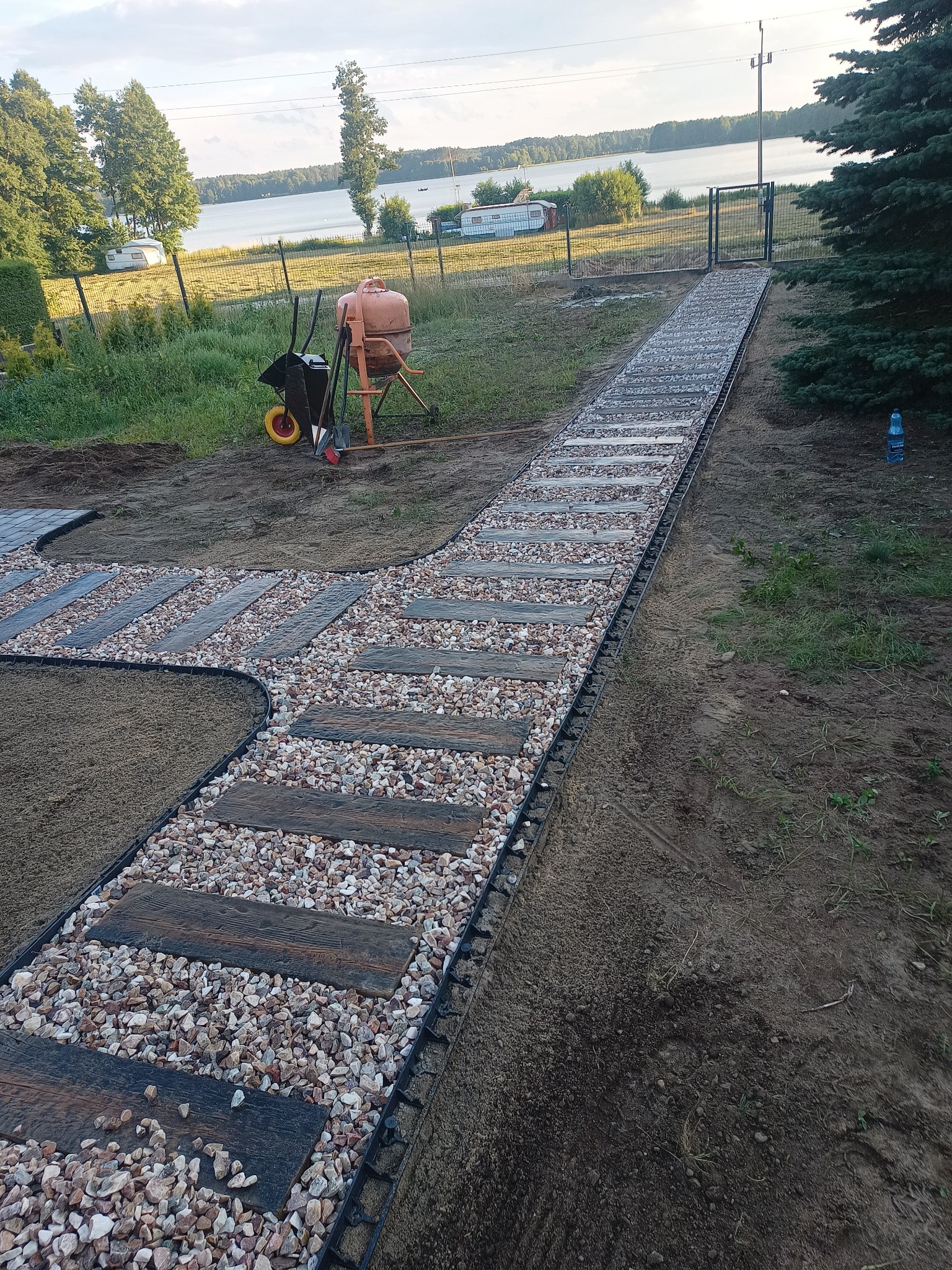 Ścieżka w ogrodzie z drewnianych elementów i jasnych kamieni, prowadząca do bramy, w tle jezioro i mikser do betonu.