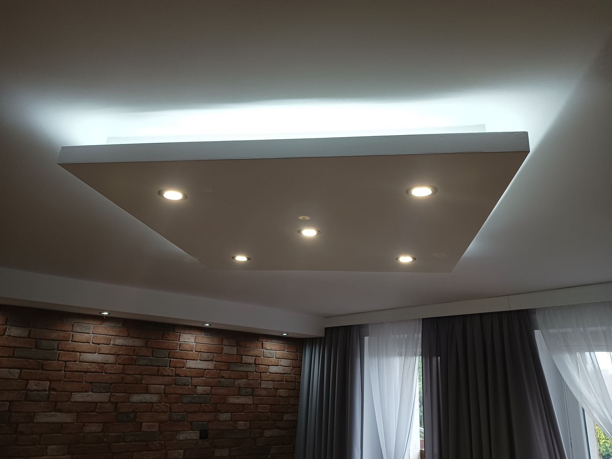Podwieszany sufit z oświetleniem punktowym w kwadratowej wnęce, widok z dołu na tle ściany z cegły i zasłon.