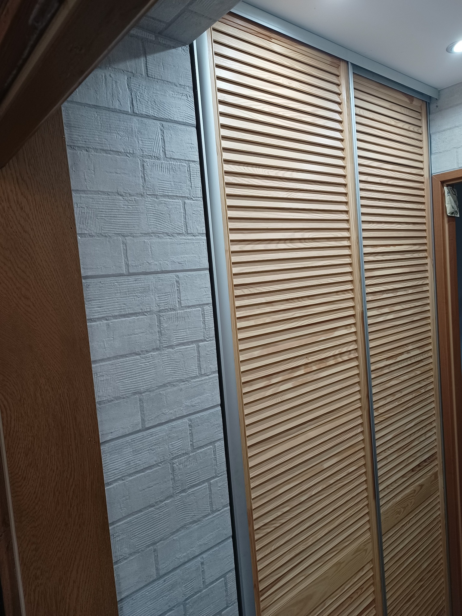 Dwie przesuwne drewniane żaluzjowe drzwi szafy wnękowej z metalowymi prowadnicami, na tle ściany z białej cegły strukturalnej i fragmentu drewnianej framugi drzwi.