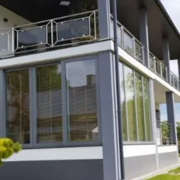 Szare okna z żaluzjami w nowoczesnym domu z balkonem z metalową balustradą i szklanymi panelami, widok z zewnątrz na trawnik.