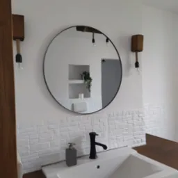 Nowoczesna łazienka z okrągłym lustrem, białymi płytkami imitującymi cegłę, czarną armaturą i drewnianymi akcentami oświetleniowymi.