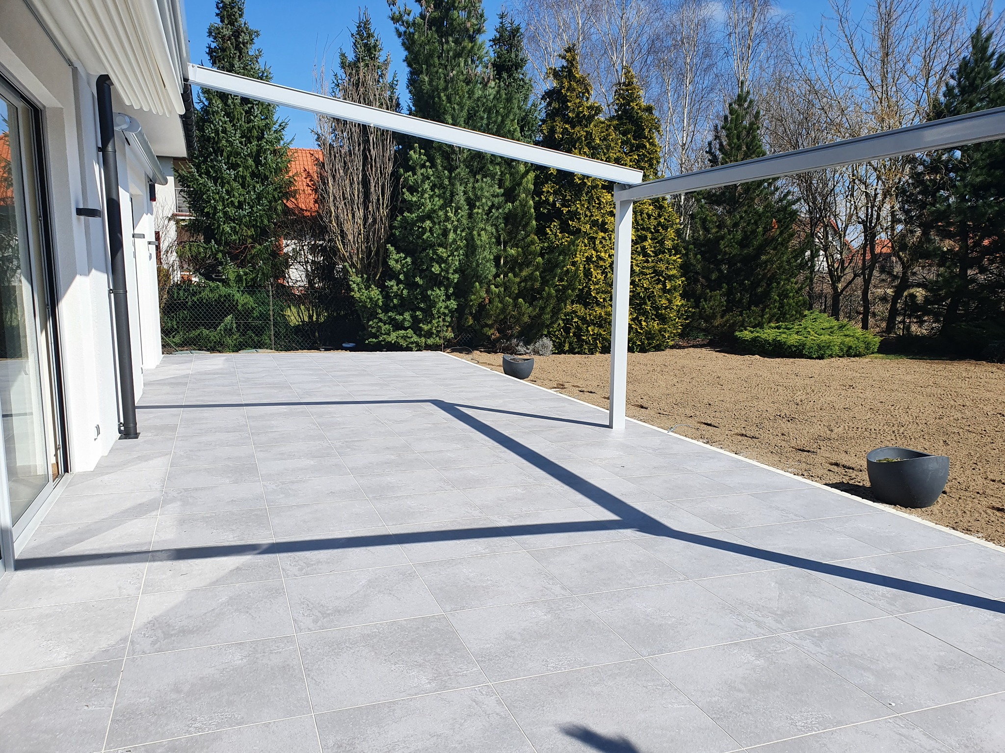 Nowoczesny taras z szarych, kwadratowych płyt kamiennych, zadaszony białą pergolą, z widokiem na ogród z drzewami iglastymi i dwoma donicami w tle.