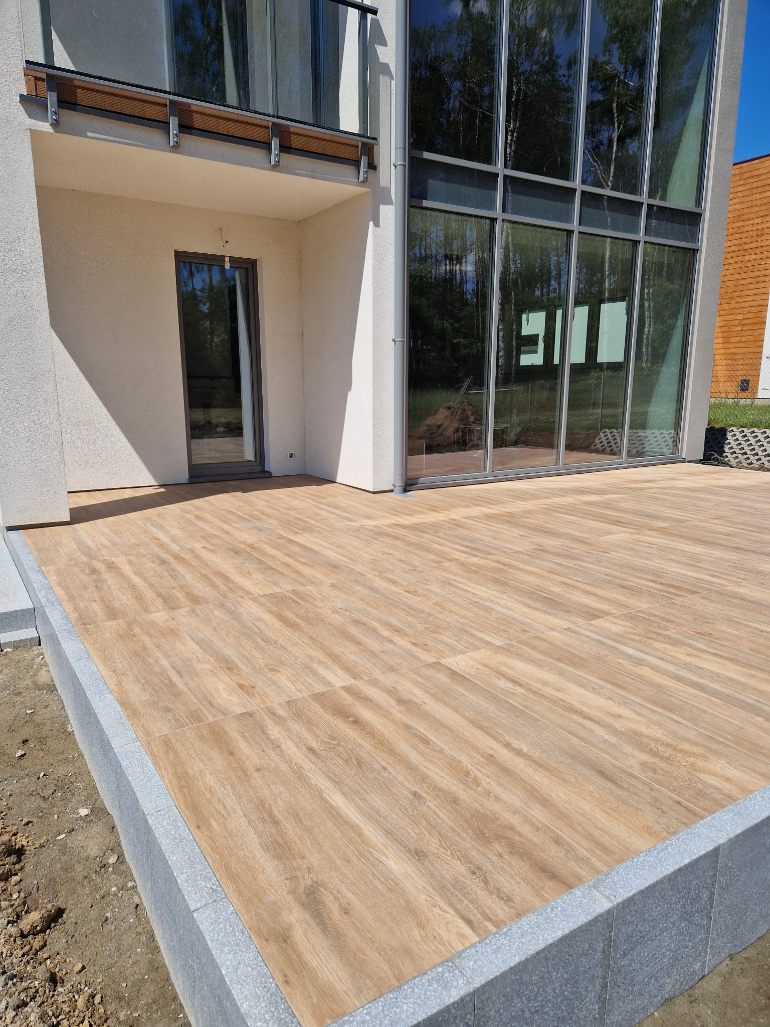 Nowoczesny taras z płytek imitujących drewno, obrzeże z szarego granitu, duże okna domu z odbiciem zieleni.