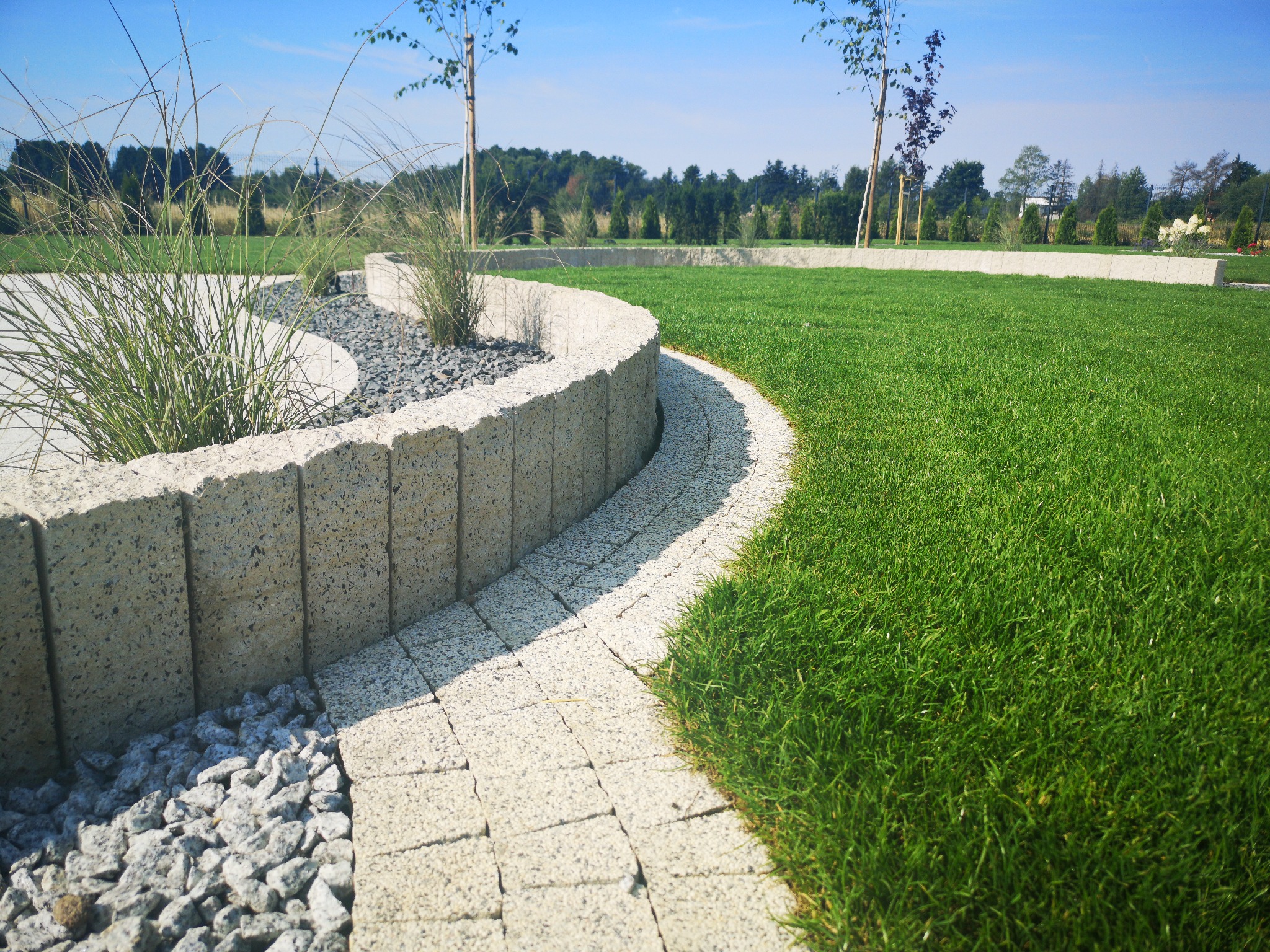 Zakrzywiony chodnik z kostki brukowej prowadzi przez trawnik, obok niskiego murku z roślinami ozdobnymi i kamieniami dekoracyjnymi.