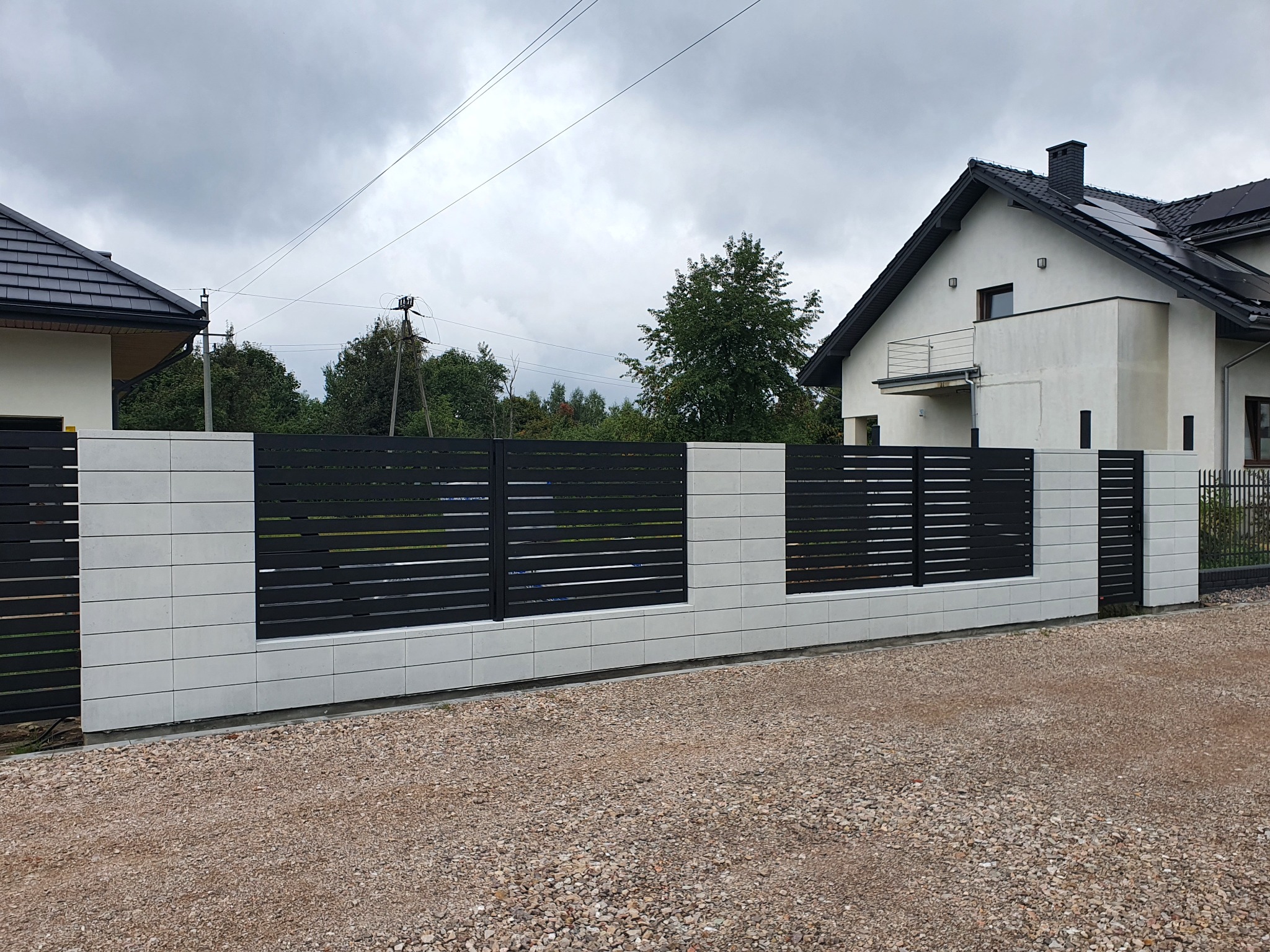 Nowoczesne ogrodzenie z poziomych, czarnych paneli między segmentami z jasnoszarych, betonowych bloczków, na tle domu z panelami słonecznymi.