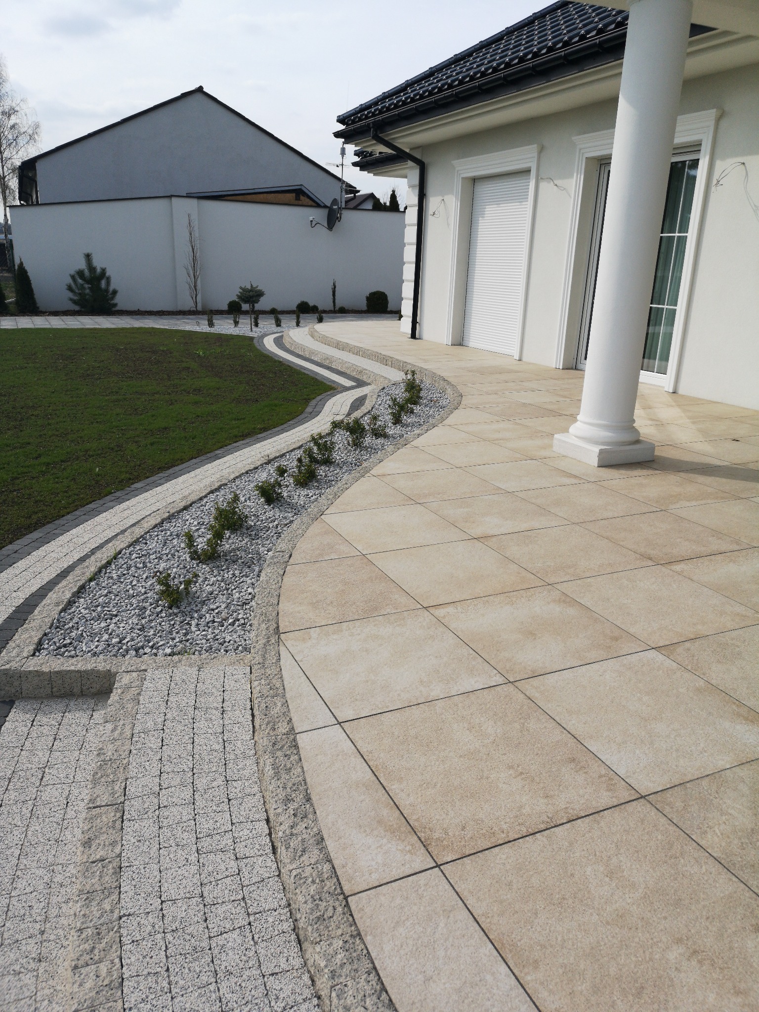 Nowoczesny taras z jasnych, kwadratowych płyt, obok ścieżka z kostki brukowej i rabata z białym żwirem oraz niskimi krzewami, dom z kolumną w tle.