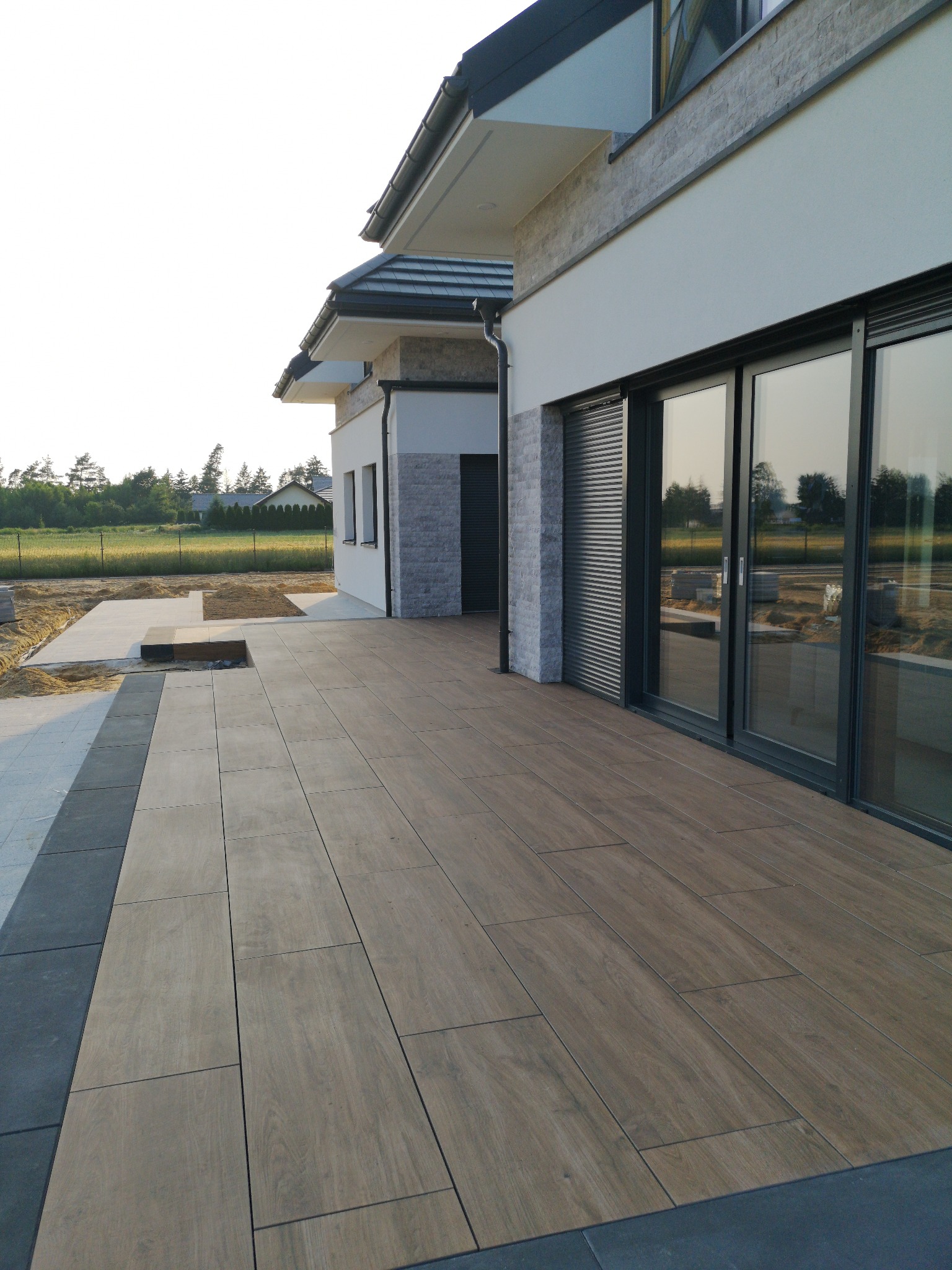 Nowoczesny taras z desek kompozytowych w Konstantynowie Łódzkim, przylegający do domu z dużymi przesuwnymi oknami, z widocznym fragmentem przygotowywanej nawierzchni z kostki obok.
