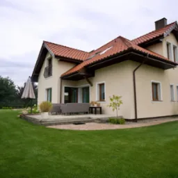 Dwupiętrowy dom z czerwonym dachem, beżową elewacją i zadbanym trawnikiem, widoczny z boku, z tarasem i meblami ogrodowymi.