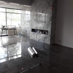 M-BUD Sp. z o.o. - Polerowana, ciemna posadzka przemysłowa z widocznym odbiciem okna i drzew, marmurowa ściana z wnęką oraz narzędzia i materiały budowlane w trakcie prac wykończeniowych.