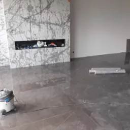 Błyszcząca, ciemnoszara posadzka z efektem marmuru, częściowo pokryta kurzem po remoncie, z odkurzaczem w lewym dolnym rogu i fragmentem ściany z marmurowym wykończeniem w tle.