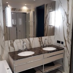 M-BUD Sp. z o.o. - Nowoczesna łazienka z dwoma umywalkami na marmurowym blacie, podświetlane lustro na ścianie z imitacją marmuru, widoczna wanna i kabina prysznicowa w tle.